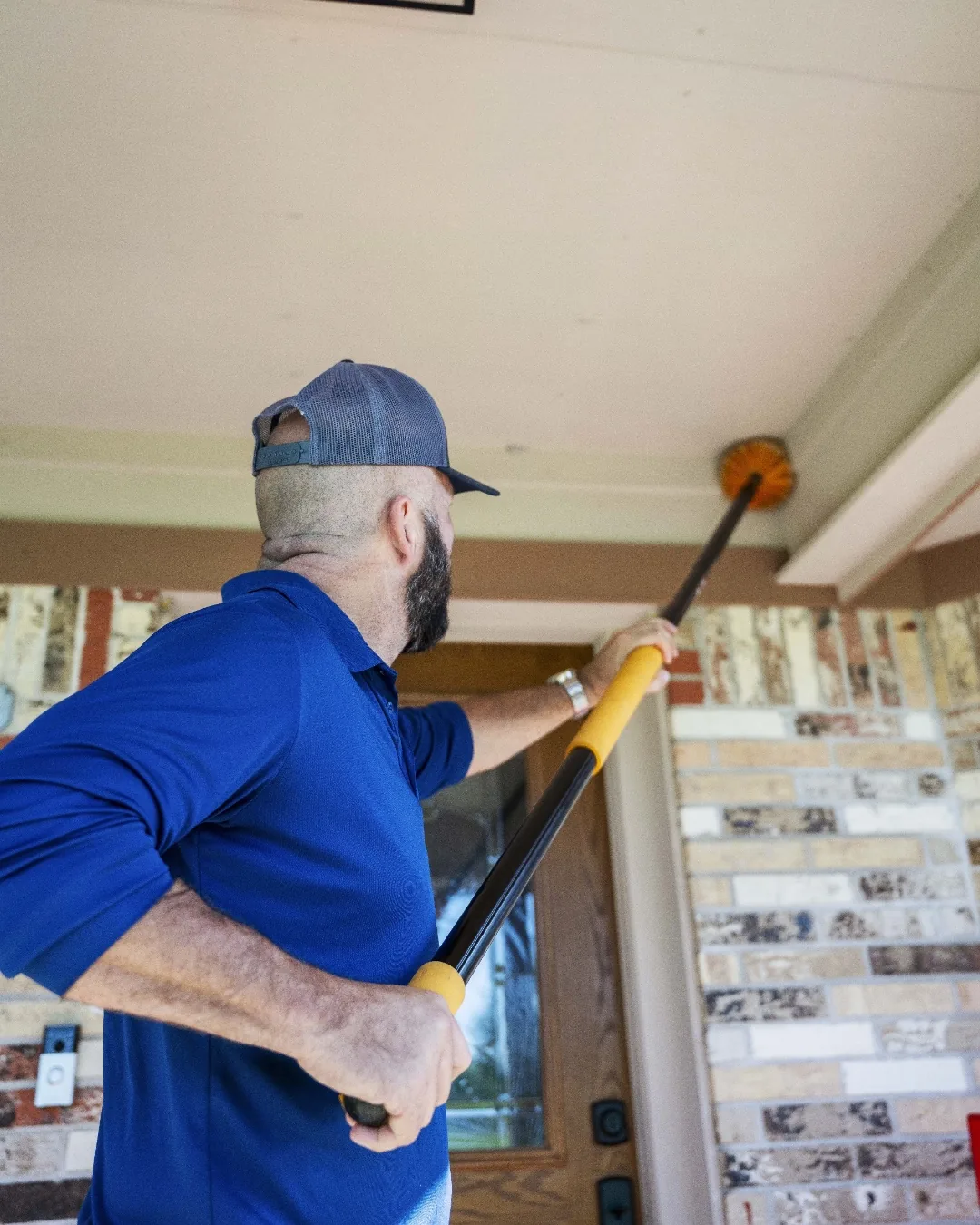 A Spot On technician removing spider webs from the porch soffit of a brick East Texas home