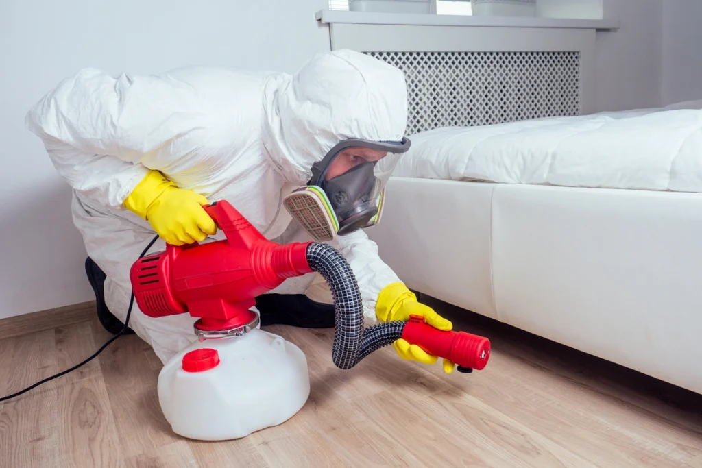Bed Bug Exterminator in Lufkin, TX 1 A man in a white suit and yellow mask sprays a bed with a vacuum for cleaning or pest control.